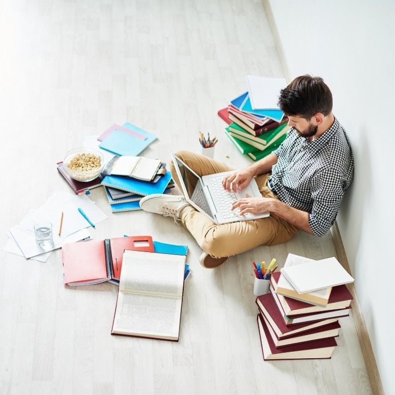 A man sits on the floor typing on a laptop, surrounded by stacks of books, capm exam practice questions, colored pencils, and a bowl of popcorn. He appears focused, working in a bright, tidy space.