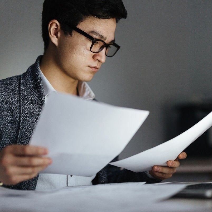 A person wearing glasses and a gray blazer sits at a desk, looking intently at documents—perhaps reviewing results after failing the CAPM exam. The blurred background keeps focus on their determined expression and work.
