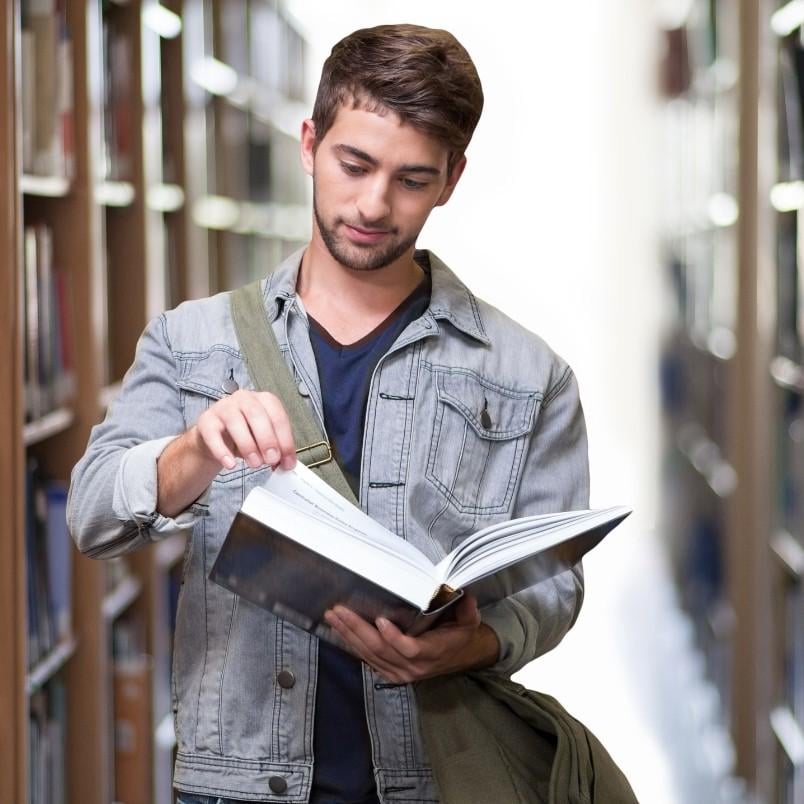 A young man with a beard, wearing a gray denim jacket and a green shoulder bag, stands in a library aisle, reading and flipping through the pages of an open book filled with CAPM exam practice questions.