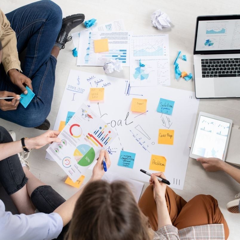 Three people sit on the floor around charts, graphs, and sticky notes, discussing business strategies and release instructions. A laptop and tablet display data, with crumpled papers scattered around. The word "Goal" is written at the center.