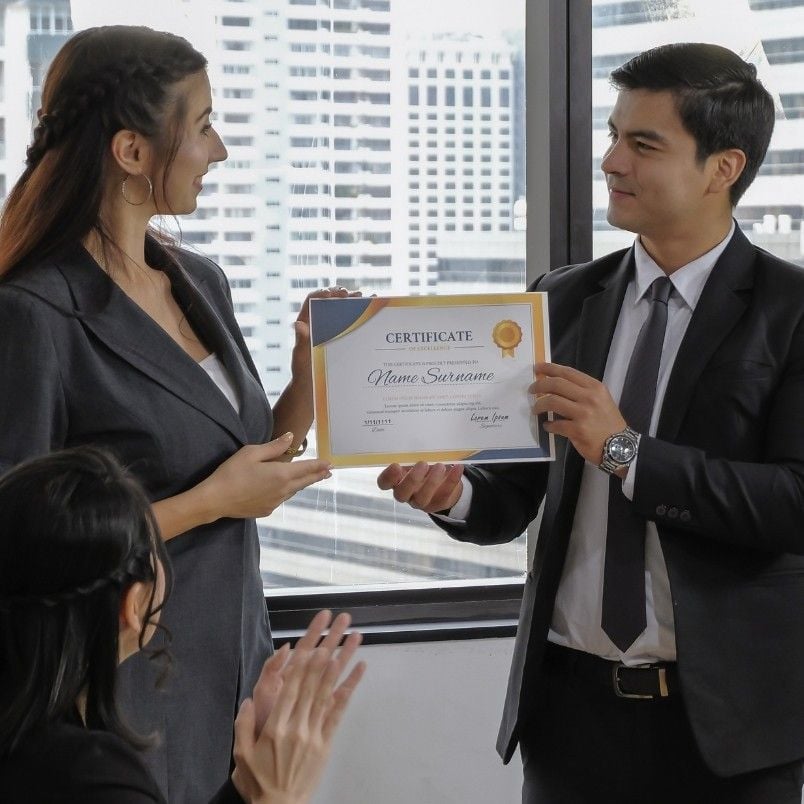 A woman in business attire receives a certificate from a man in a suit—perhaps for mastering how to pass the CAPM exam—while another person applauds in an office with large windows and city buildings in the background.