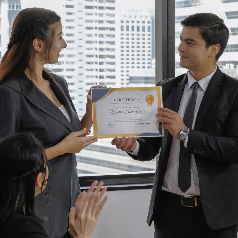A man in a suit hands a CAPM Certification Worth It certificate to a woman in business attire while another person nearby applauds. They stand before large windows with city buildings visible outside.
