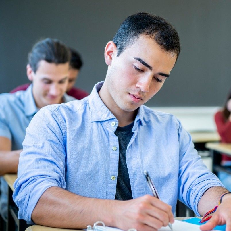 A young man in a blue shirt sits at a classroom desk, focused on writing notes about CAPM exam domains. Other students are visible in the background, also working, with a chalkboard behind them.