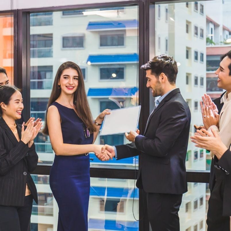 A woman in a blue dress shakes hands with a man in a suit while receiving a certificate, celebrating her achievement of meeting PMP certification requirements. Three colleagues clap and smile nearby in a modern office with large windows.