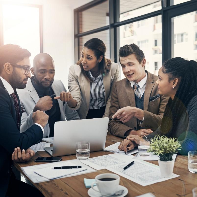 Five business professionals gathered around a table, engaged in project communication while looking at a laptop. Papers, glasses of water, and a coffee cup are on the table. The group appears focused and collaborative in a modern office setting.