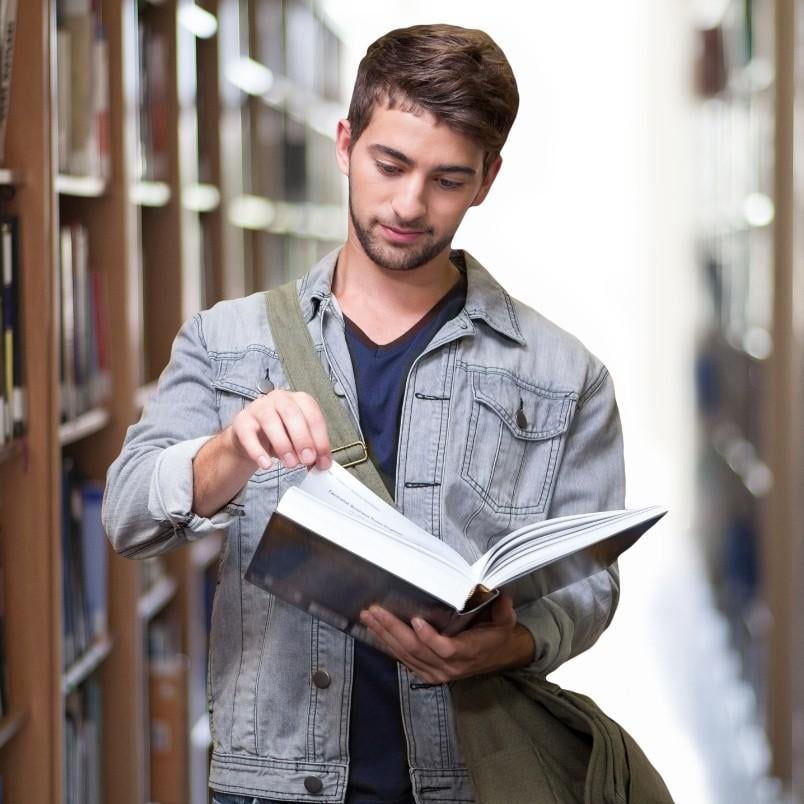 A young man with a denim jacket and shoulder bag stands in a library aisle, looking down as he flips through the pages of an open CAPM certification book. Shelves filled with books line both sides of the aisle.