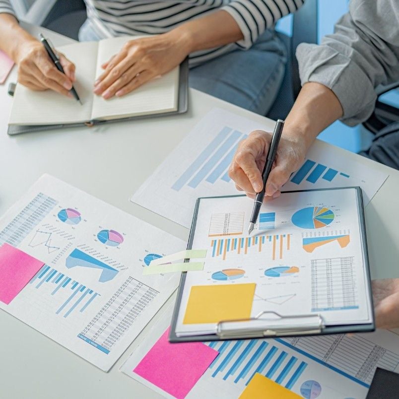 Two people are working at a desk with reports, charts, and graphs. One writes in a notebook while the other points at a clipboard full of colorful business charts, sticky notes, and uses checklist analysis for better organization.