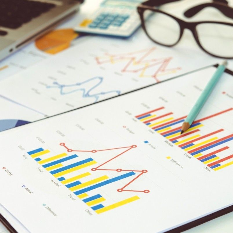 Close-up of printed financial charts with colorful bar and line graphs, a pencil resting on the paper, eyeglasses, a calculator, and a laptop—ideal for Root Cause Analysis of financial trends.