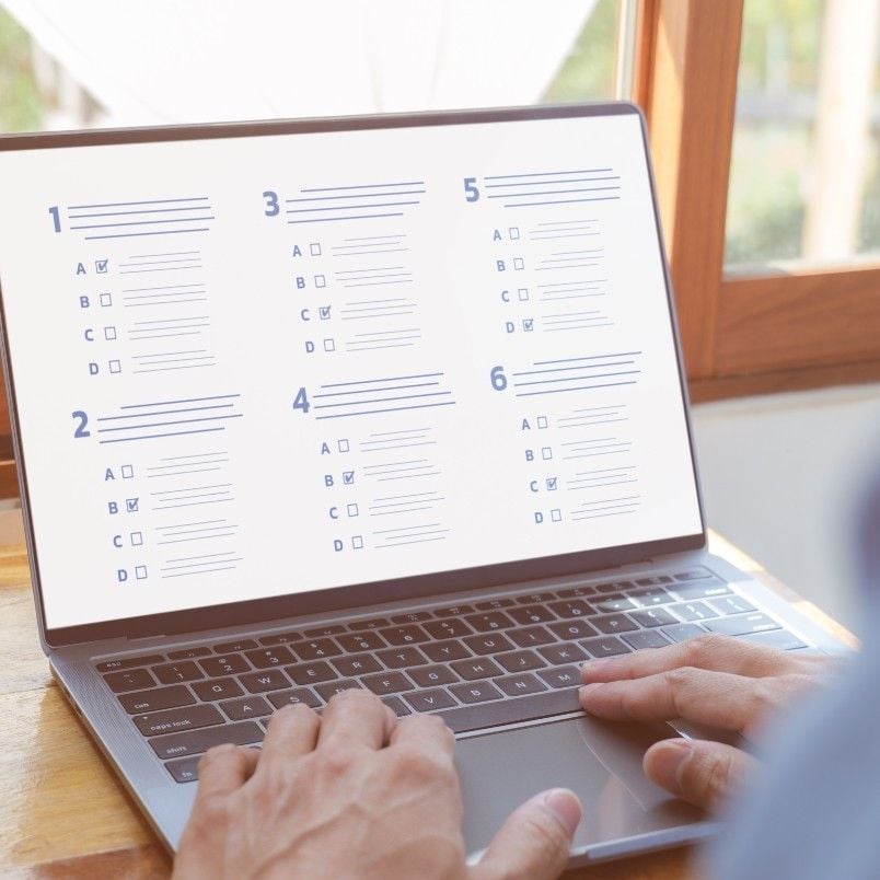 A person uses a laptop displaying a CAPM Exam Simulation with an online multiple-choice test of six questions. Some answers are checked, while sunlight streams in through the window in the background.