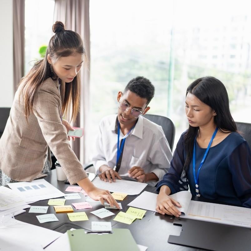 Three people in a modern office collaborate on a procurement management plan at a table covered with documents and colorful sticky notes. One woman stands and points to notes, while the other two sit and listen attentively.