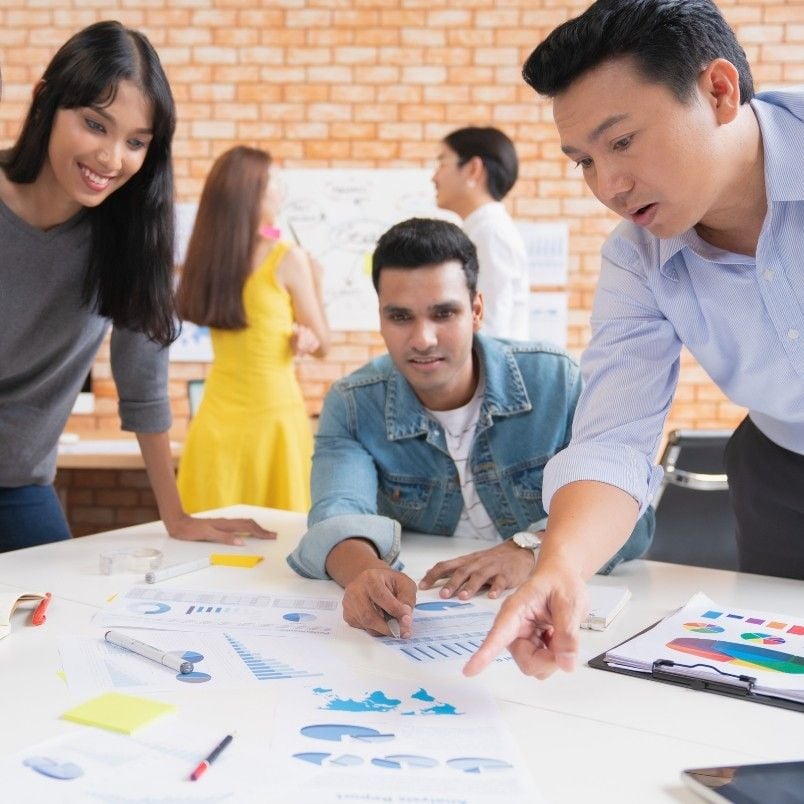 Three people gather around a table covered with charts and graphs, discussing data and product roadmap instructions. Two others converse in the background near a brick wall covered with papers and diagrams.