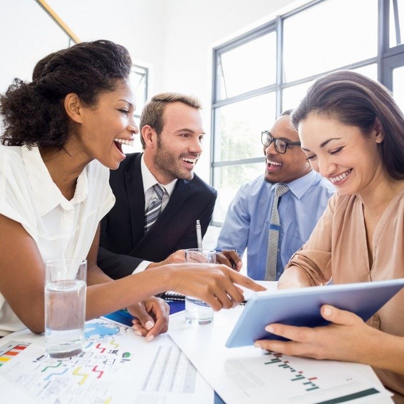 Four smiling colleagues sit at a table with charts and glasses of water, reviewing data and Retrospective Instructions together in a bright, modern office. They appear engaged as they collaborate around a tablet.
