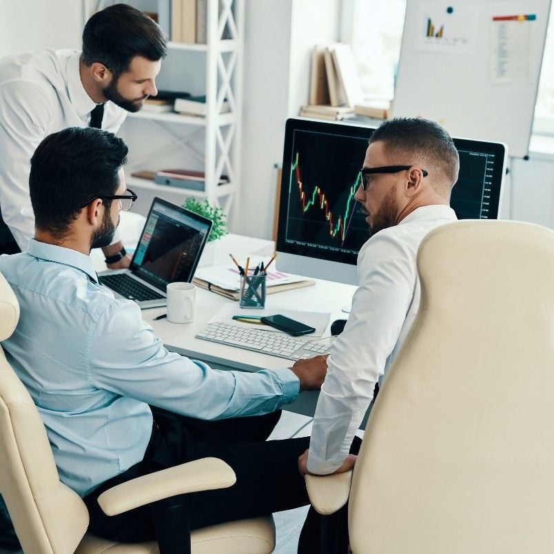 Three businessmen in an office work at computers displaying financial charts, discussing data and reviewing a risk management plan. One stands and leans over while the other two sit at desks, focusing on the monitors.