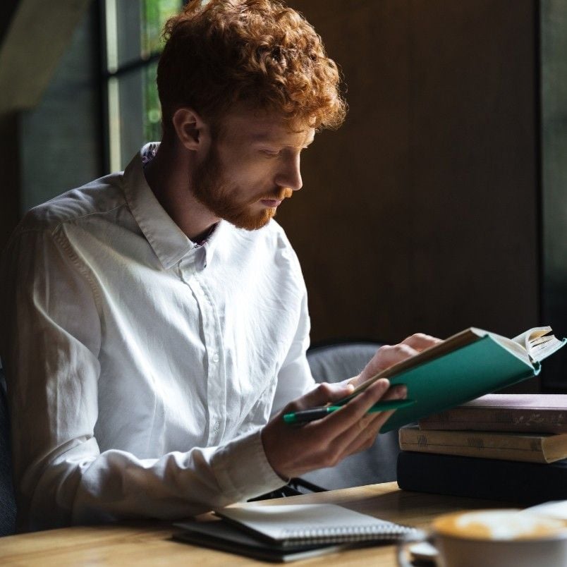 A person with curly red hair and a white shirt sits at a table, studying hard after failing the CAPM exam. Other books and a notebook are on the table, and soft light streams in from a nearby window.