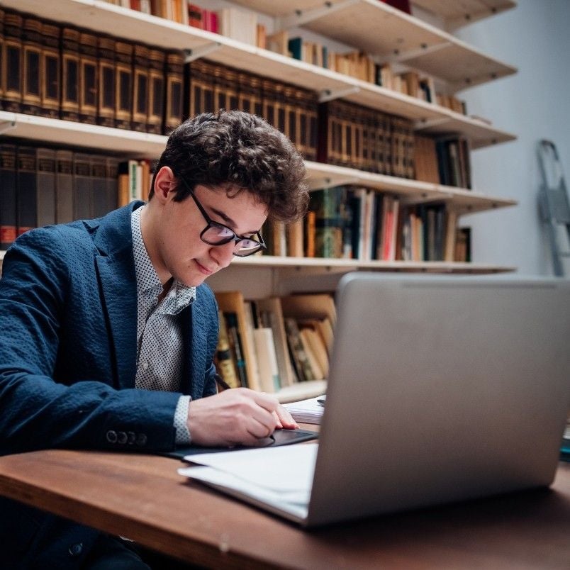 A young man wearing glasses and a blazer sits at a desk in a library, preparing for the CAPM Exam Simulation by reading a book and using his laptop. Shelves filled with books line the background.