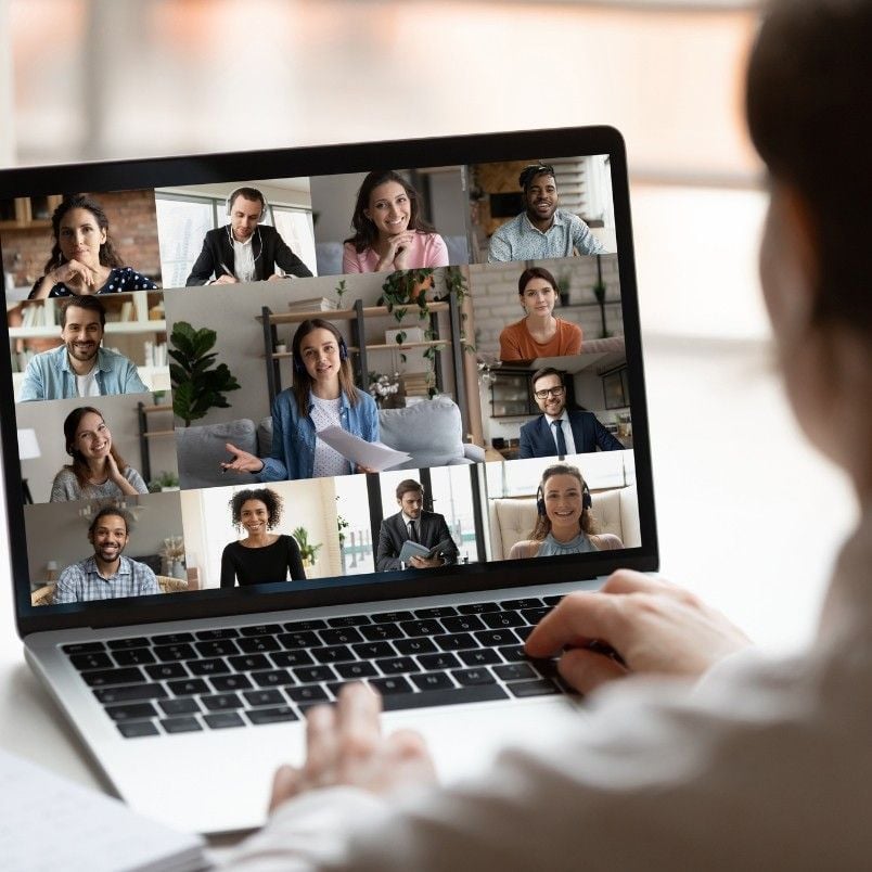 A person participates in a video conference on a laptop, with twelve diverse people shown in a grid on the screen, perhaps discussing strategies after failing CAPM exam, each appearing engaged from individual locations.