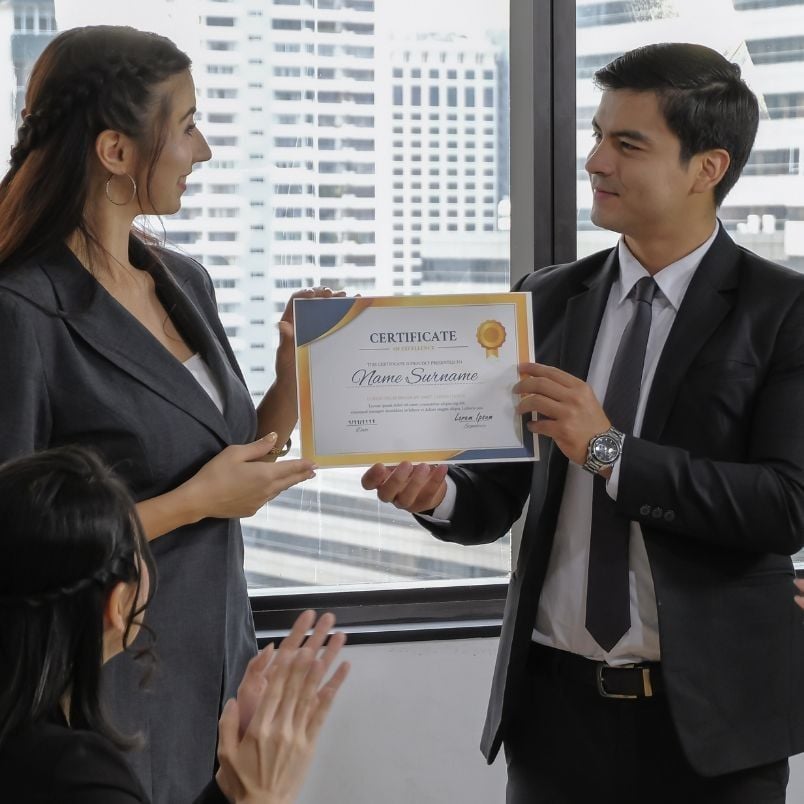 A woman in a suit hands a certificate to a man in a suit as another person applauds, celebrating the achievement of PMP certification requirements. They stand before large windows with city buildings visible in the background.