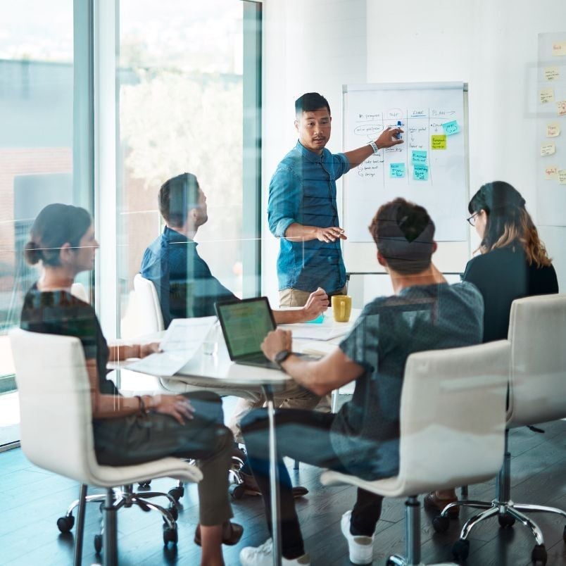 Five people are in a meeting room. One person stands by a whiteboard covered with charts about PMP certification requirements, presenting to four colleagues who are listening attentively and taking notes on laptops and notepads around the table.