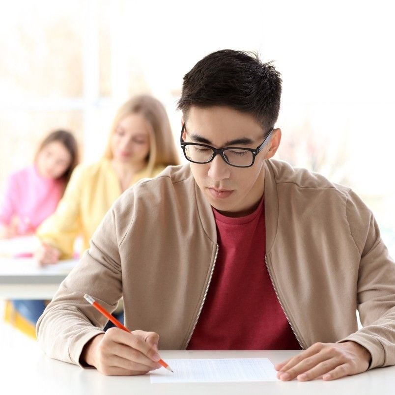 A young man wearing glasses and a beige jacket writes with a pencil at a desk, focusing on capm exam practice questions. Two other students sit in the blurred background.