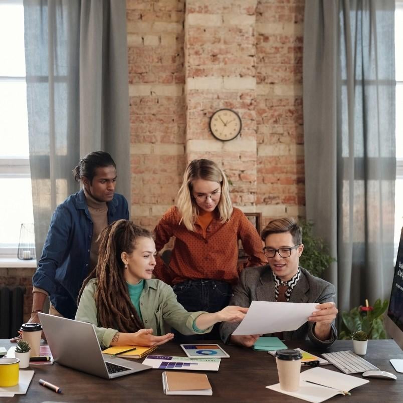 Four people gather around a desk covered with laptops, notebooks, and coffee cups, reviewing product roadmap instructions together in a modern office with brick walls and large windows.