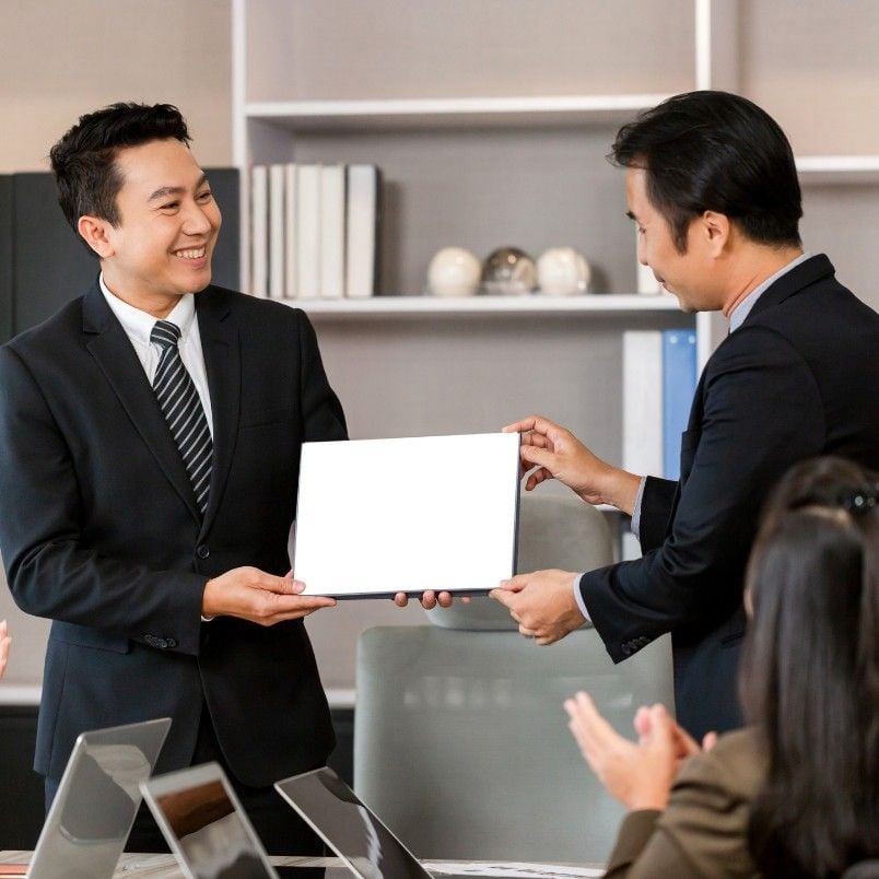 Two men in business suits smile as one hands a blank certificate to the other in an office setting, while people in the foreground clap—celebrating success, perhaps after learning how to pass CAPM exam together.