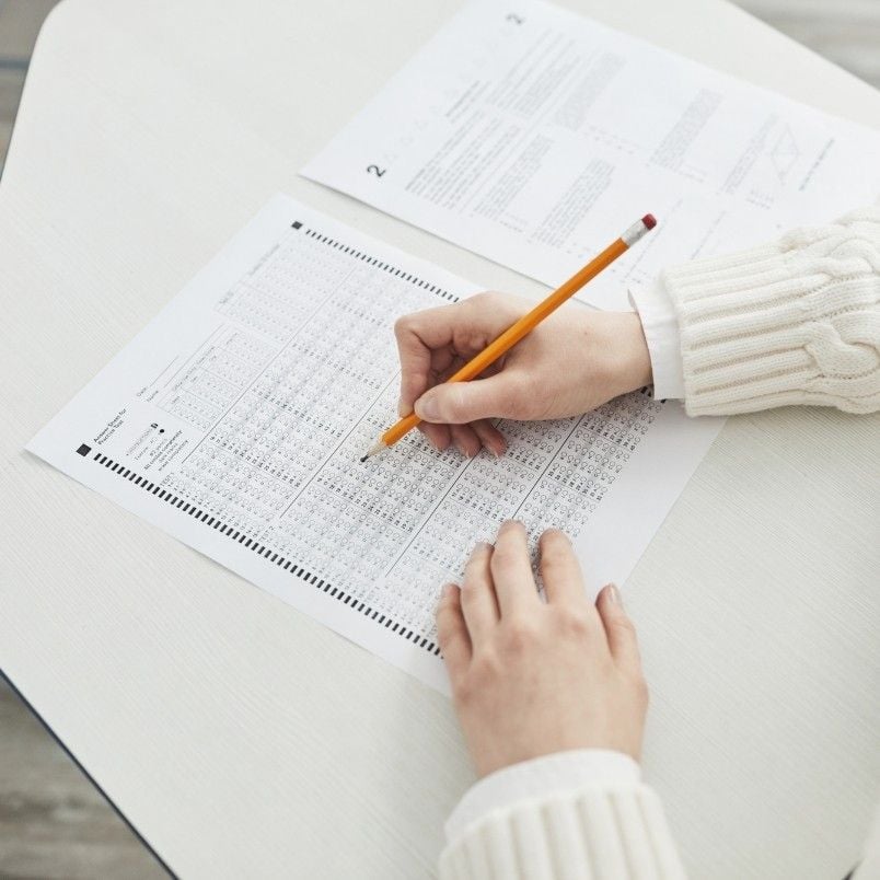 A person wearing a white sweater is filling out capm exam practice questions on a multiple-choice answer sheet with a pencil on a desk, with a test booklet open nearby.