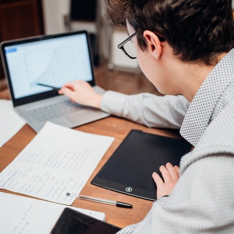 A person sits at a desk analyzing a line graph on a laptop. Papers with handwritten notes, capm exam practice questions, a pen, a tablet, and a smartphone are also on the desk. The person is focused on the computer screen.