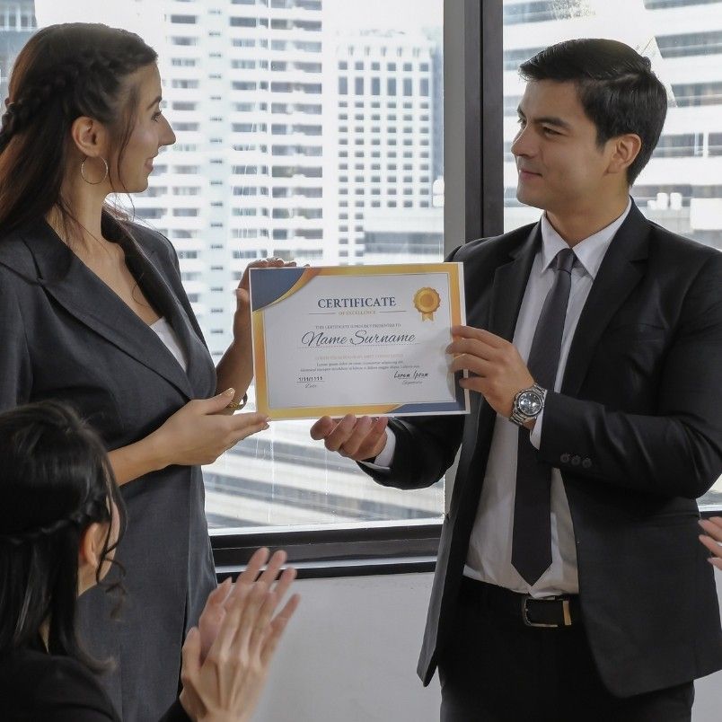 A woman in business attire receives a certificate for completing her CAPM certification training from a man in a suit while colleagues applaud in a modern office with large windows and city buildings in the background.