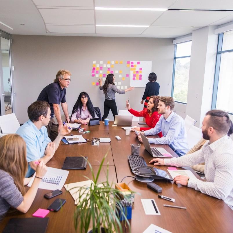 Eight people collaborate in a modern conference room, some using laptops and papers while others stand by a whiteboard covered in colorful sticky notes, applying the nominal group technique to discuss and prioritize ideas.