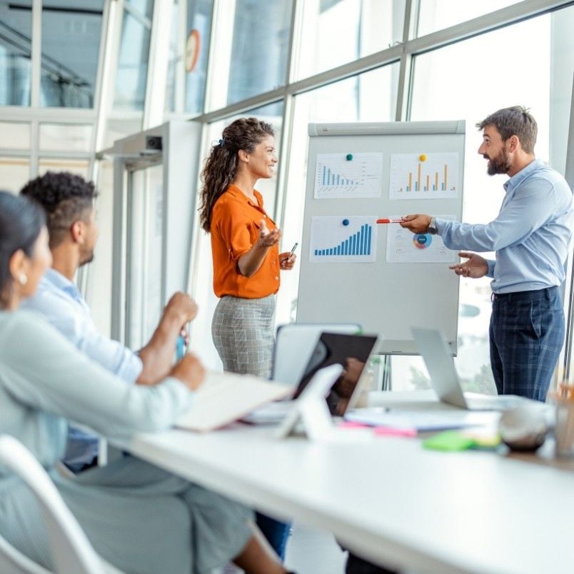 Four people have a business meeting in a bright office. Two stand by a flipchart with graphs, discussing product roadmap instructions, while two others sit at the table with laptops and notebooks, listening and watching the presentation.