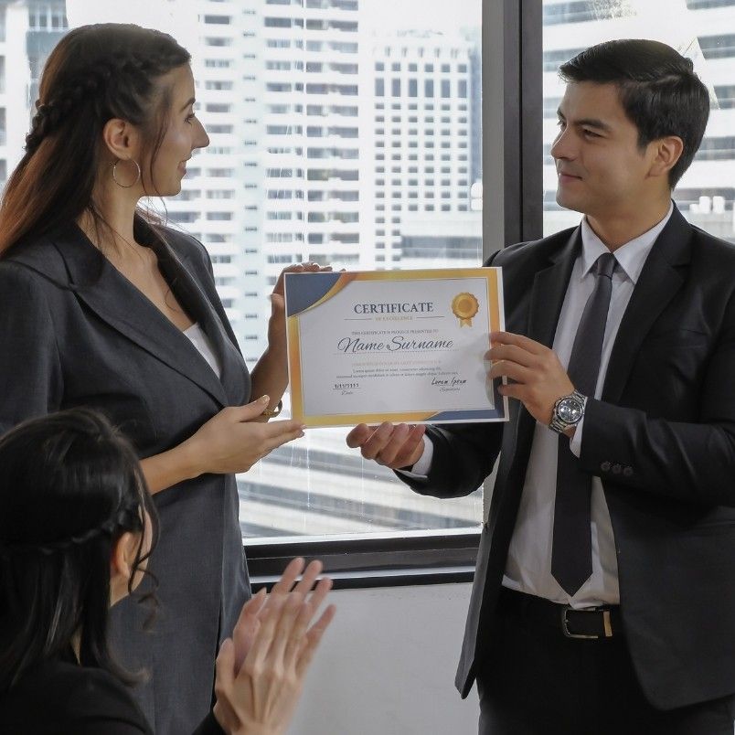 A man in a suit hands a certificate to a woman in business attire—perhaps for mastering CAPM exam domains—while another person applauds in the foreground. They are indoors with large windows overlooking city buildings.