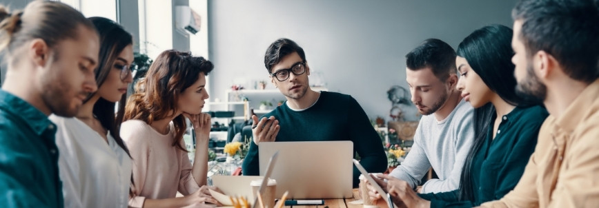 A group of seven young adults sit around a table in an office, engaged in a serious discussion about feasibility assessment instructions. Laptops, notebooks, and papers are visible as they focus on a man in glasses who appears to be leading the conversation.