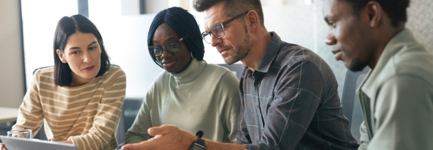 Four people sit together at a table, looking at a laptop screen and discussing a change management plan. They appear focused and engaged in a collaborative work or study setting.