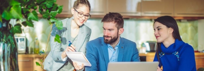 Three people sit at a table with a laptop. One woman stands, smiling as she writes on a notepad and reviews a checklist, while the man and another woman beside him listen attentively in a bright, modern office space.