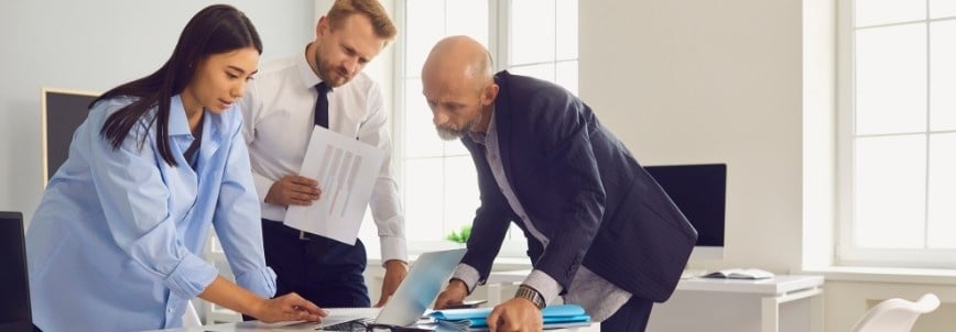 Three business professionals, two men and one woman, are standing around a desk in a bright office, reviewing documents and discussing how to use a decision tree to guide their work together.