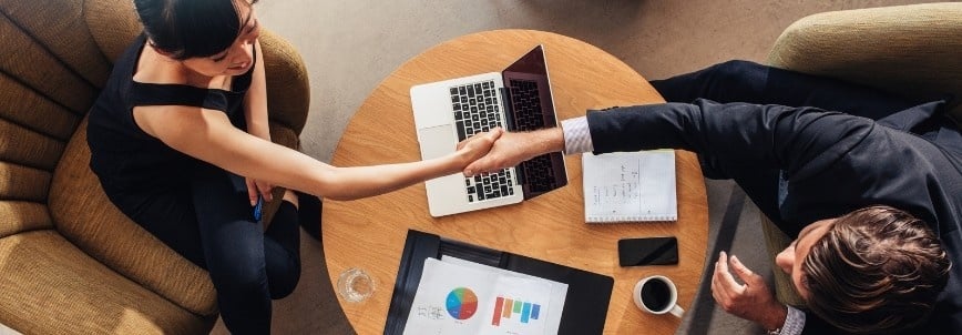Two people sit across a small round table, shaking hands. A laptop, documents with colorful charts, a notebook, and a glass of water suggest a business meeting or project handover.