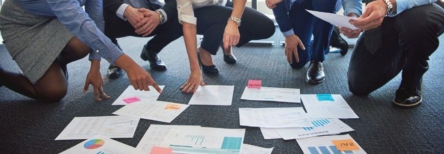 Four people in business attire kneel on the floor, examining charts, graphs, and documents spread out on dark carpet, using mind mapping techniques with colorful sticky notes among the papers.