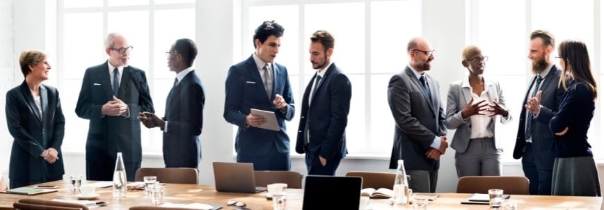 A group of eight professionals in business attire stand and talk around a long conference table with laptops, papers, and water bottles, resembling a focus groups session in a bright, modern office with large windows.