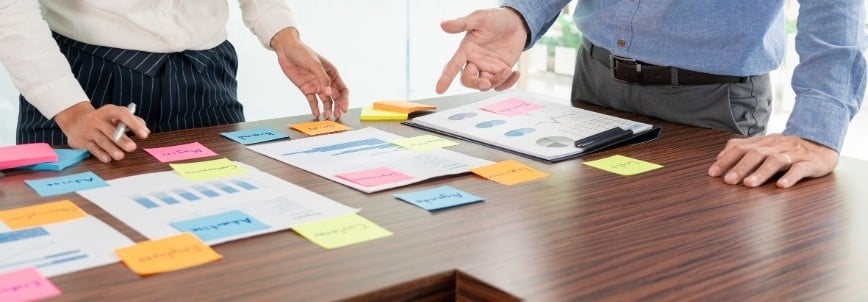 Two people stand at a table covered with reports, charts, and colorful sticky notes, discussing and organizing business documents such as the Quality Management Plan during a meeting.