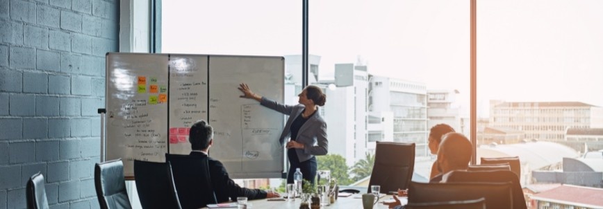A person stands at a whiteboard presenting a resource management plan to three seated colleagues in a modern conference room with large windows overlooking a cityscape.