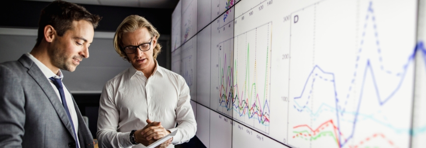Two men in business attire discuss cost-benefit data in front of large screens displaying various colorful line graphs and charts in a modern office setting.