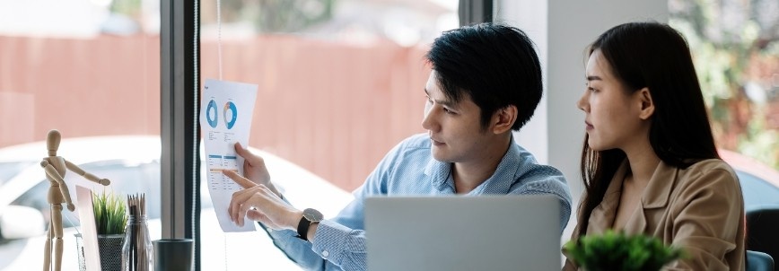 A man and woman sit at a desk by a window, reviewing a paper with charts and graphs on earned value analysis. The man points to the document while the woman listens. A laptop and office supplies are on the table.
