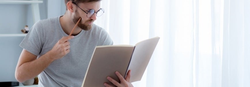 A man wearing glasses and a gray t-shirt sits indoors, holding a pencil to his lips and reading PMP study materials in a large book, appearing thoughtful. Light filters through sheer curtains behind him.