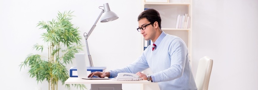 A man wearing glasses and a light blue sweater sits at a white desk, focused on drafting a procurement statement of work on his computer in a bright office with a lamp, files, and a potted plant in the background.