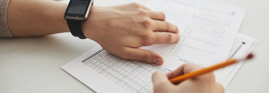 A person wearing a smartwatch holds a test paper, while another hand fills in answers with a pencil on a multiple-choice answer sheet with CAPM practice questions.
