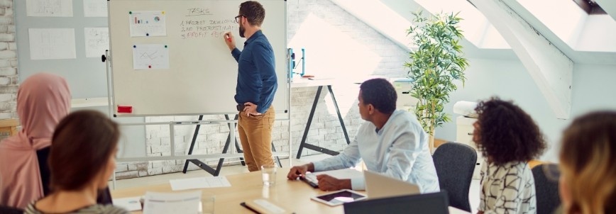 A man stands at a whiteboard writing while four colleagues, seated at a conference table, watch and listen attentively as he discusses project management and performance reporting in a bright, modern office space.