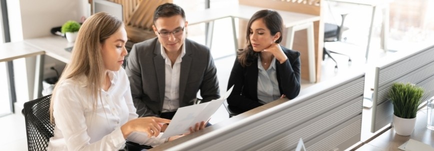 Three people in business attire sit at desks in a modern office, discussing a team charter that one woman is holding and pointing at, while the others attentively listen.