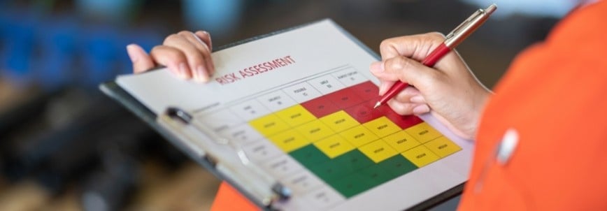 Person holding a clipboard with an agile risk register template, marking a section on the color-coded risk assessment chart—green, yellow, and red—with a red pen. The individual is wearing an orange sleeve.