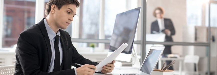 A young man in a suit sits at a desk in a modern office, reviewing documents—perhaps preparing for pmp exam situational questions—while looking at his computer. In the background, another person stands by a window holding papers.