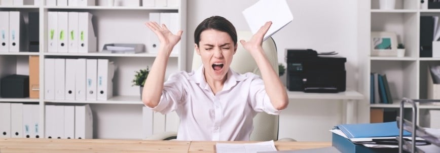 A frustrated woman sits at a desk in an office, raising her arms and shouting while holding papers labeled with Risk Adjusted Backlog Instructions, surrounded by shelves filled with binders and documents.